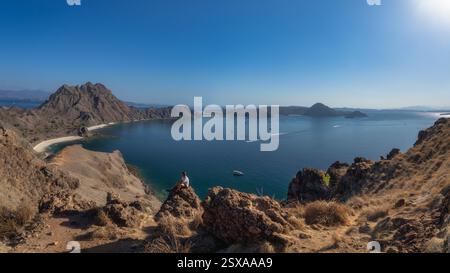 Ein atemberaubender Panoramablick zeigt einen Mann, der auf einem Felsen sitzt, eine ruhige Küste mit kristallklarem Wasser und zerklüftetem felsigem Gelände von Pada Stockfoto