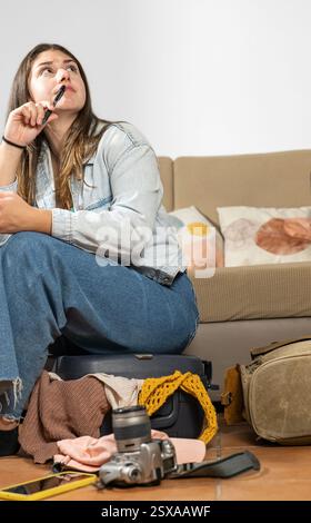 Eine junge Frau, die auf ihrem kleinen Koffer sitzt und mit einem Bleistift in der Hand ihre Reiseriste durchsucht. Konzentration, während sie prüft, ob alles bereit ist Stockfoto
