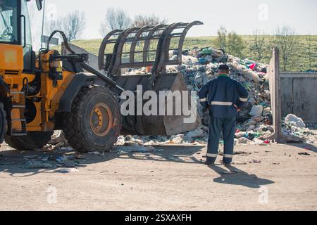 Ein Arbeiter im Recycling-Center, in dunkelblauer Arbeitskleidung, der sich einen unsortierten Müllhaufen ansieht, ist gerade in der Materialrückgewinnungsanlage angekommen, Rückansicht. Stockfoto