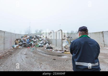 Ein Arbeiter im Recycling-Center, in dunkelblauer Arbeitskleidung, der sich einen unsortierten Müllhaufen ansieht, ist gerade in der Materialrückgewinnungsanlage angekommen, Rückansicht. Stockfoto