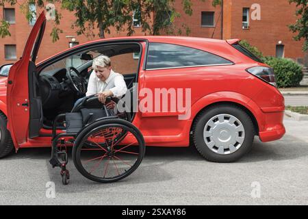Lächelnde Frau mit Behinderung sitzt auf dem Fahrersitz und hebt ihren Rollstuhl in das Auto, auf den Beifahrersitz. Stockfoto