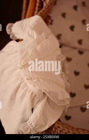 Ein weißes Babykleid für das Kleinkind hängt an der beigen Babywiege im Schlafzimmer, gemütliches Gefühl wie zu Hause in der Familie, Familie schätzt den Lebensstil. Stockfoto