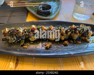 Köstliche knusprige Tempura-Uramaki-Sushi-Brötchen werden mit Aalsauce, Sesamsamen und gehacktem Schnittlauchschnitt garniert, serviert auf einem schwarzen Serviergericht Stockfoto