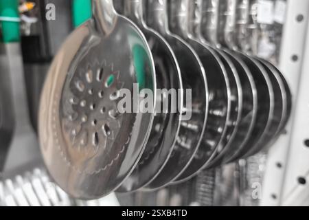 Verschiedene Küchenutensilien aus Metall hängen an einem Ausstellungsregal in einem hellen kulinarischen Laden. Die polierten Oberflächen reflektieren das Licht und zeigen ihre Handwerkskunst. Stockfoto
