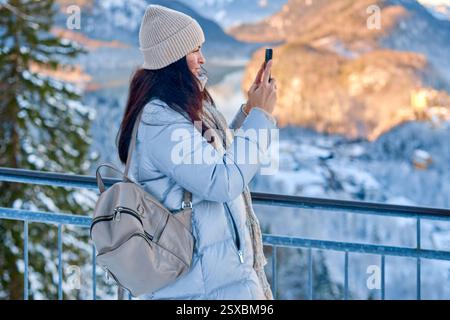 Dieses hochauflösende Foto zeigt eine ruhige Winterszene mit einer jungen Frau, die ihr Smartphone in einer schneebedeckten Berglandschaft nutzt. Stockfoto