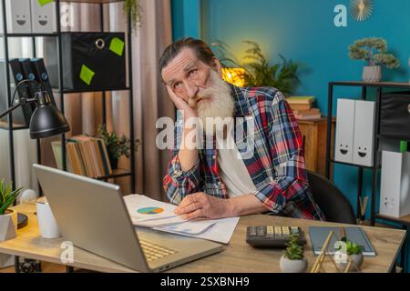 Müde erschöpfter Senior Old man Freelancer, der Papierkramme analysiert, Zahlungen abrechnet und monatliche Ausgaben Bankrechnungen am Home Office Desk berechnet. Verärgerter Opa, der am Tisch arbeitet. Stockfoto