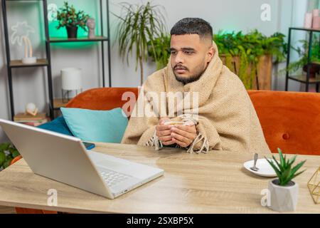 Kranker, kranker indischer Mann, der an einem Laptop arbeitet, der in Karo gewickelt ist, sitzt zitternd vor kaltem trinken heißem Kaffee im unbeheizten Heimbüro, ohne Schulden zu heizen. Der ungesunde Hindu-Typ versucht sich aufzuwärmen Stockfoto