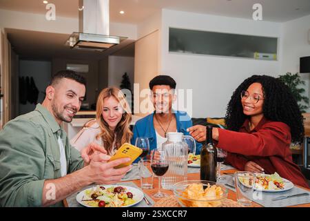 Multirassische Freunde genießen ein köstliches Abendessen zusammen, während sie Lachen teilen und Momente mit einem Smartphone in einem gemütlichen Essbereich voller köstlicher Speisen und freudiger Gespräche festhalten. Hochwertige Fotos Stockfoto