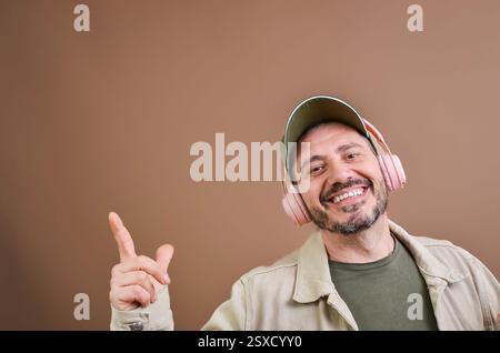 Lächelnder Mann mit Kopfhörern und Baseballmütze, der den Finger auf braunem Hintergrund zeigt Stockfoto