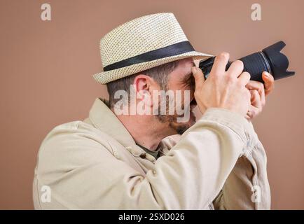 Lächelnder Fotograf mit beigefarbenem Hut, der Fotos mit professioneller Kamera im Studio mit braunem Hintergrund macht Stockfoto