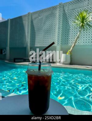 Eiskaffee am Pool ein Eiskaffee in einer Plastiktasse mit Strohhalm steht auf einem weißen Tisch mit einem türkisfarbenen Pool im Hintergrund. Stockfoto