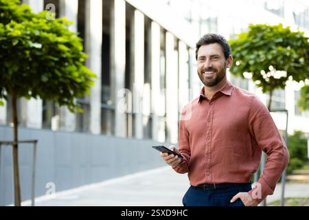 Porträt eines erfolgreichen und freudigen Geschäftsmannes. Ein Mann mit einem Telefon in der Hand lächelt und schaut in die Kamera, während er aus einem Bürogebäude geht. Stockfoto