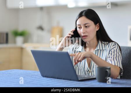 Asiatische Frau, die am Telefon über Laptop-Inhalte in der Küche zu Hause spricht Stockfoto