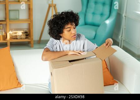 Afrikanisches Mädchen, das die Lieferung auspackt und in die Schachtel schaut. Glückliche Frau, die eine Schachtel öffnet. Eine Frau bekommt ein Paket, das sich die gelieferten Waren ansieht. Zufriedene Kunden positives Feedback, schnelle Expresslieferung Stockfoto