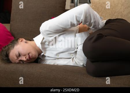 Geschäftsfrau mit weißem Hemd und dunkler Hose liegt auf einem Sofa, hält ihren Magen vor Schmerzen, ihr Gesicht drückt Unbehagen und Leiden aus Stockfoto