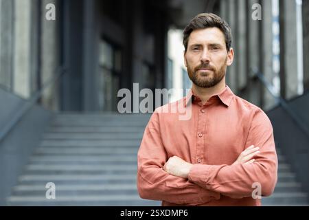 Porträt eines ernsthaften selbstbewussten Geschäftsmannes vor einem Bürogebäude, Mann mit überkreuzten Armen, der selbstbewusst in die Kamera blickt Stockfoto