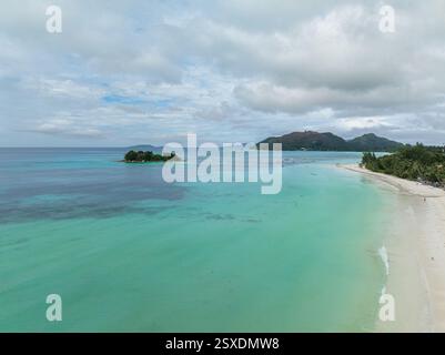 Blick aus der Vogelperspektive auf türkisfarbenes Wasser, das auf eine weiße Sandküste trifft, mit einer kleinen Insel in der Ferne. Anse Volbert Beach. Praslin, Seychellen. Stockfoto