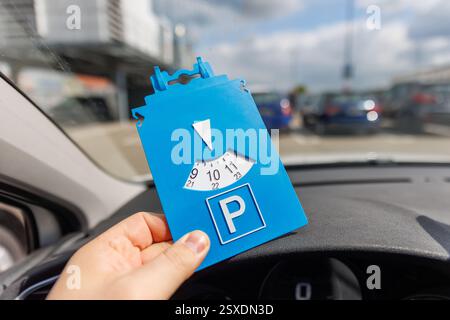 Fahrer Hand halten blaue Parkscheibe zeigt die Ankunftszeit in der Nähe eines großen Einkaufszentrums. Parkzeitbeschränkungen auf belebten Parkplätzen. Zeitlich begrenzt Stockfoto