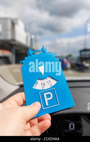 Fahrer Hand halten blaue Parkscheibe zeigt die Ankunftszeit in der Nähe eines großen Einkaufszentrums. Parkzeitbeschränkungen auf belebten Parkplätzen. Zeitlich begrenzt Stockfoto