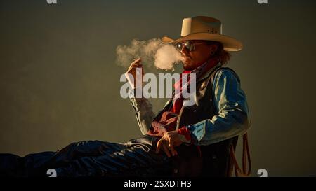 Mann als Cowboy gekleidet in Lederhosen, Jeansjacke, rauchende Zigarre, Sonnenbrille, Rauch mit selbstbewusster Einstellung. Stockfoto