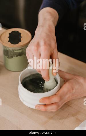 Hände, die Matcha in einer Keramikschale auf einer hölzernen Kaffeekanne rühren Stockfoto