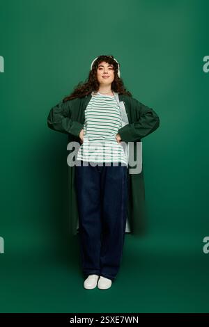 Selbstbewusste junge Frau posiert mit Händen auf den Hüften und präsentiert ihr stilvolles Outfit in einem grünen Ambiente. Stockfoto