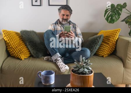 Lächelnder Mann, der das Smartphone benutzt, während er sich zu Hause auf dem Sofa entspannt Stockfoto