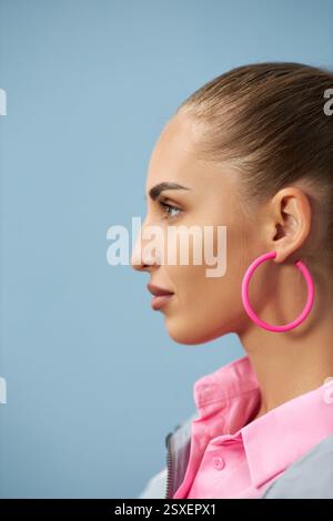 Nahaufnahme des Seitenprofils einer Frau, die wegschaut, mit großen rosa Creolen und glattem, zurückziehendem Haar vor hellblauem Hintergrund Stockfoto