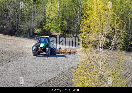 Feldbearbeitung mit Deutz-Fahr Agrotron-Traktor und Egge im Frühjahr. Landwirtschaftliche Landschaft mit Frühlingsbäumen. Salo, Finnland. Mai 2024. Stockfoto