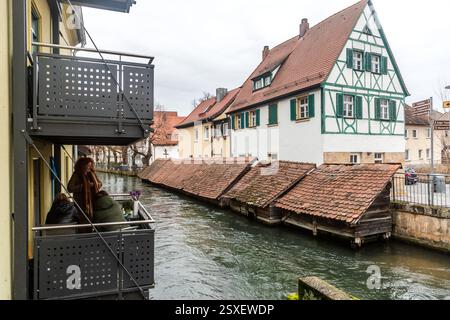 Historische Fischkästen mit moosbewachsenen Ziegeldächern liegen im Wasser der Wiesent in der Altstadt von Forchheim. Zwei Frauen sitzen im Vordergrund auf einem Balkon und blicken über die Kasten hinweg, mit einem Blumenstrauß auf dem Tisch zwischen ihnen. Im Hintergrund erhebt sich ein giebelständiges Fachwerkhaus mit grünen Fensterläden. Die hölzernen Kasten dienten Forchheims Fischhändlern seit Jahrhunderten dazu, Fische am Leben zu erhalten, nahe der historischen Hundsbrücke. Schönbornstraße, Forchheim, Bayern, Deutschland. Stockfoto