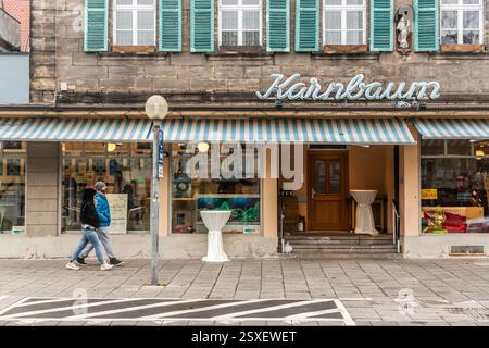 Ein Paar geht an der Fassade des Karnbaum-Fischladens in der Altstadt von Forchheim vorbei, wobei das Neonzeichen und das Aquarium im Schaufenster sichtbar sind. Vor dem Eingang steht ein Stehtisch. Das Geschäft verkauft frischen Fisch, Meeresfrüchte und Wein und gehört zu den lang etablierten Fischhändlern der Stadt. Zweibrückenstraße, Forchheim, Bayern, Deutschland. Stockfoto