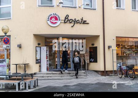 Die Bogatz-Kaffeerösterei in der Altstadt von Forchheim begrüßt zwei Fußgänger unter ihrem runden roten Logo über dem Eingang. Bänke und ein kleiner Tisch stehen vor der cremefarbenen Fassade, rechts lehnt ein Fahrrad an der Wand. Die Rösterei röstet Kaffeebohnen aus Ländern wie Brasilien, Guatemala, Indien und Äthiopien in einer traditionellen Trommelröstmaschine. Hornschuchallee, Forchheim, Bayern, Deutschland. Stockfoto