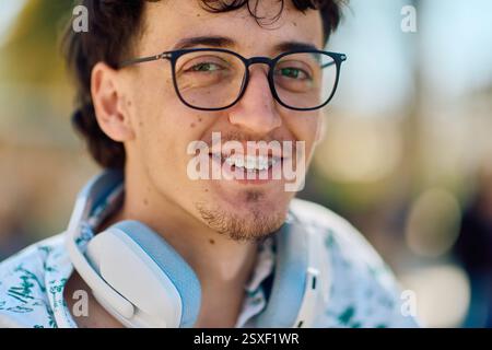 Lächelnder junger Mann mit Brille und Kopfhörern in sonniger Umgebung im Freien Stockfoto