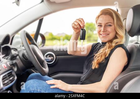 Eine lächelnde blonde Frau sitzt auf dem Fahrersitz eines modernen Autos und hält selbstbewusst die Schlüssel hoch Stockfoto