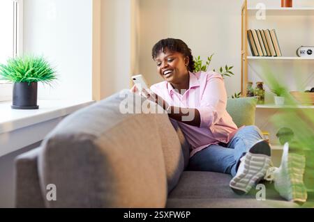 Glückliche afroamerikanische Frau zu Hause, die auf dem Sofa mit Handy sitzt, um sich mit einem Freund zu unterhalten Stockfoto