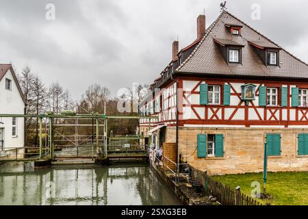 Ein historisches Fachwerkhaus mit einer rot-weißen Fassade, grünen Fensterläden und einer Wetterfahne auf dem Dach steht neben einer Schleusenanlage an der Wiesent in Kirchehrenbach. Eine Heiligengestalt sitzt in einer Nische zwischen den Fenstern, mit einem schlichten weißen Gebäude, das links angrenzt. Der grün gestrichene Schleusenrahmen spannt sich über den Wasserlauf im Vordergrund. Weilersbacher Straße, Kirchehrenbach (VGem), Bayern, Deutschland. Stockfoto