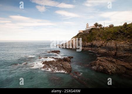 Ein malerischer Leuchtturm steht auf einer zerklüfteten, von grüner Vegetation bedeckten Klippe entlang der Küste von Cudillero, Asturien, Spanien Stockfoto
