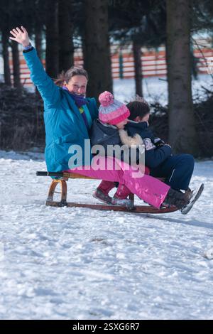 Eine lachende und winkende Frau rutscht mit zwei Kindern auf einem Schlitten einen schneebedeckten Hügel hinunter Stockfoto