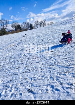 Zwei Kinder auf kleinen Schlitten rutschen einen steilen, verschneiten Hügel hinunter Stockfoto