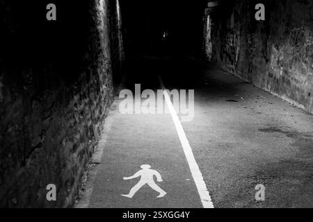 Ein Fußgängerweg, der mit einer weißen Linie und einem Symbol für eine Wanderfigur markiert ist, führt in eine dunkle und enge Gasse, die auf eine ausgewiesene Route hinweist. Stockfoto