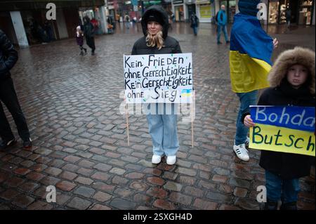 Flensburg, Schleswig-Holstein Person mit Transparent in der Hand mit der Aufschrift kein Frieden ohne Gerdchtigkeit,. Keine Gerechtigkeit ohne Sieg. Der Überfall Russlands auf die Ukraine jährt sich am 24. Februar 2025 zum dritten Mal. Aus diesem Anlass findet statt in Flensburg auf dem Südermarkt eine Kundgebung. Viele Kinder, eine erdrückende Stimmung, die meien Menschen aus der Ukraine, Menschen die fliehen müssen, auf dem Boden liegen Spielsachen, Plüschtiere, Fähnchen, Kerzen. Mit einem roten Band wird das Land Ukraine nachgebildet. Aufnahme vom 24.02.2025, Flensburg *** Flensburg, Schle Stockfoto