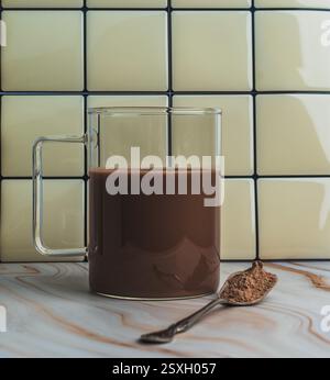 Ein klarer Glasbecher mit reichhaltiger heißer Schokolade steht auf einer Marmorarbeitsfläche neben einem kleinen Löffel Kakaopulver vor einer gefliesten Wand. Stockfoto