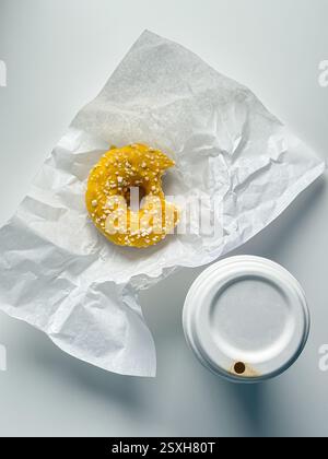 Gebissener gelber Donut in weißem Papier auf weißem Bürotisch mit Kaffeetasse zum Mitnehmen Stockfoto