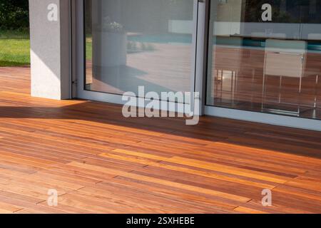 Hartholzboden auf der Terrasse neben einem Haus mit einem großen Glasfenster. Natürliches Licht unterstreicht die satten Farbtöne einer Holzterrasse außerhalb eines Contempo Stockfoto