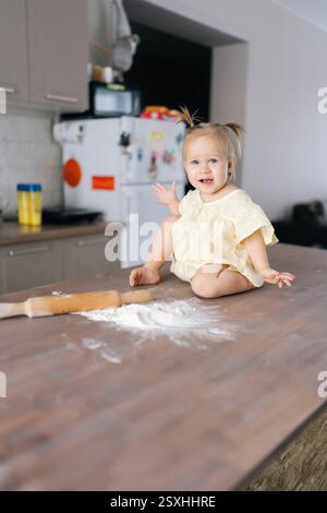 Vertikales Porträt eines fröhlichen Kleinkindes, das mit Mehl spielt, das auf dem Küchentisch sitzt, hölzerne Nadel neben ihr Stockfoto