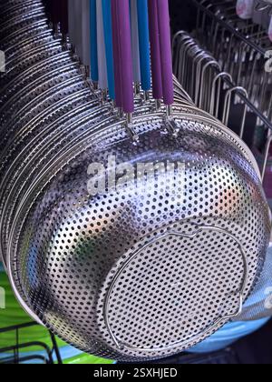 Im Sonnenlicht leuchten Metallsiebe, die von einem Regal unter farbenfrohen Küchenutensilien aufgehängt werden. Der geschäftige Markt ist lebendig mit Käufern, die verschiedene h Stockfoto