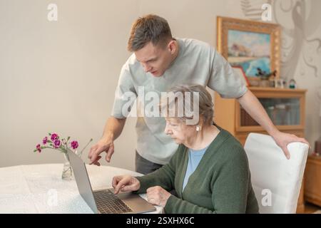 Junger Mann, der einer älteren Frau hilft, einen Laptop zu Hause zu benutzen. Konzept von Technologie, Bildung und digitaler Anpassung. Enkel unterrichtet Stockfoto