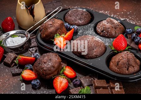 Schokoladen-Muffins mit Beeren. Muffin-Pfanne mit hausgemachten süßen, dunklen Schokoladen-Cupcakes mit Kakaosoße, Zuckerpulver und frischen Erdbeeren, Blaubeeren Stockfoto