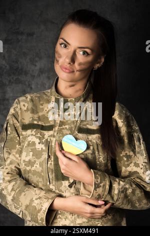 Schöne junge Militärfrau in Militäruniform mit Tarnschminke im Gesicht hält eine herzförmige ukrainische Flagge in den Händen. Studiofoto o Stockfoto