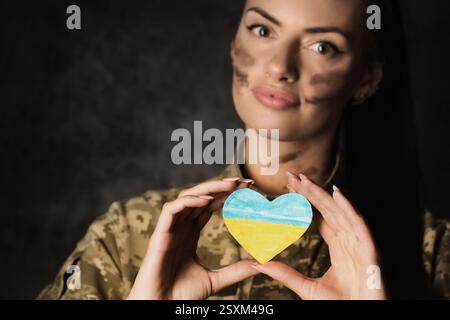 Schöne junge Militärfrau in Militäruniform mit Tarnschminke im Gesicht hält eine herzförmige ukrainische Flagge in den Händen. Studiofoto o Stockfoto